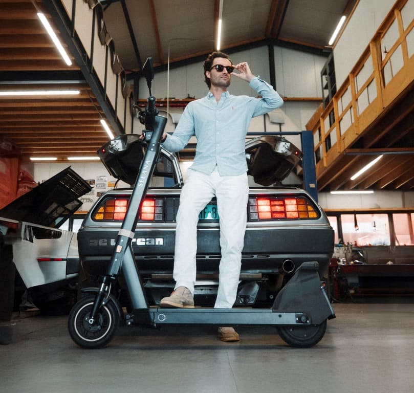 Iemand naast een e-step in een werkplaats, met een klassieke sportwagen op de achtergrond.